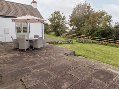 An outdoor patio with a stone floor a round table with six chairs and an umbrella beside a white house with green doors and a garden with trees and a wooden fence at Fountain Folly in Moylgrove near St Dogmaels