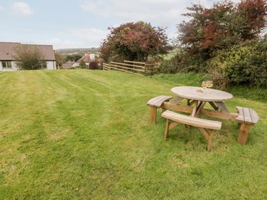 A wooden picnic table with benches on a lawn with houses trees and a fence in the background at Fountain Folly in Moylgrove near St Dogmaels