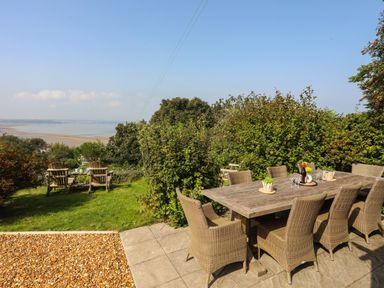 An outdoor dining area with a large wooden table and wicker chairs on a patio near a grassy area with wooden chairs and a view of the water at Gorphwysfa in Llanddona near Beaumaris