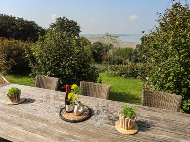 An outdoor wooden table with wine bottle glassware and plants on a patio overlooking a garden and a distant beach at Gorphwysfa in Llanddona near Beaumaris