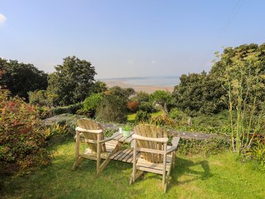 Two wooden chairs with a small table on grass overlooking trees and a distant beach at Gorphwysfa in Llanddona near Beaumaris