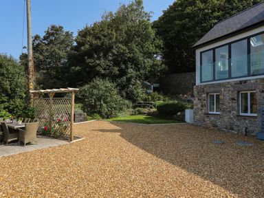 An outdoor patio dining area with chairs and table next to a gravel driveway and a stone house with large windows and trees in the background at Gorphwysfa in Llanddona near Beaumaris