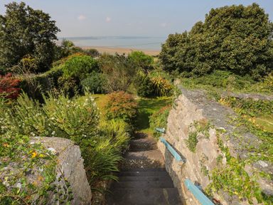 Stone steps with blue handrails leading down to a garden with trees and shrubs and a view of the beach and sea at Gorphwysfa in Llanddona near Beaumaris