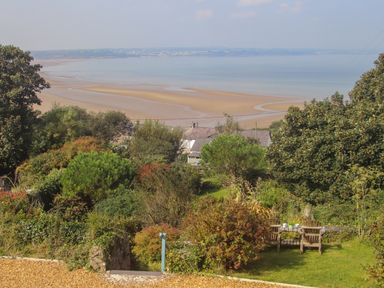 A garden with trees and shrubs overlooking a sandy beach and sea with houses in the distance at Gorphwysfa in Llanddona near Beaumaris