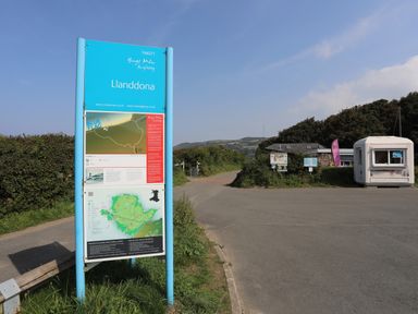 A blue information sign with maps and text beside a road near bushes and a white kiosk at Gorphwysfa in Llanddona near Beaumaris