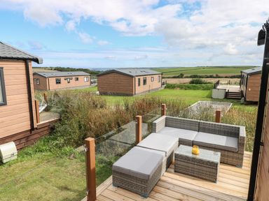 An outdoor patio with a wicker sofa and table surrounded by glass panels overlooking wooden cabins and green fields at Sandy Bay Retreat Hasguard Cross near Broad Haven
