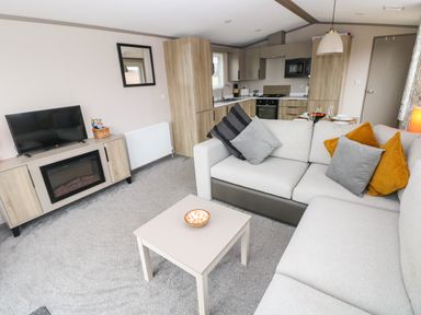 A living room with a gray sofa and cushions a small table with a bowl a TV on a wooden stand and a kitchen in the background at Sandy Bay Retreat in Hasguard Cross near Broad Haven