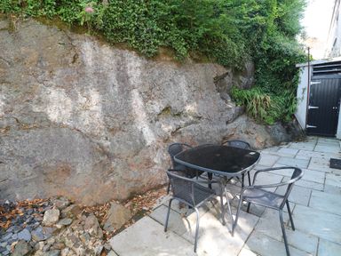 A patio with a black table and four chairs next to a rock wall and plants at 8 Rosewall Cottages in St Ives