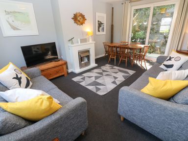 A living room with two grey sofas with pillows a geometric rug a wooden dining table with chairs a fireplace a flat screen TV and a sliding glass door at 8 Rosewall Cottages in St Ives