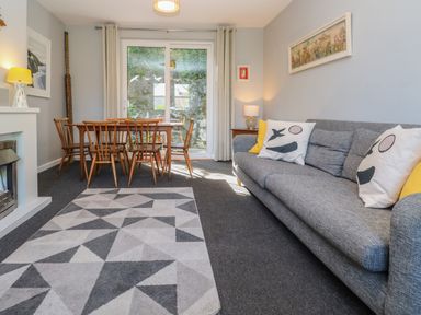 A living room with a gray sofa with cushions a patterned rug a wooden dining table with chairs and sliding glass doors at 8 Rosewall Cottages in St Ives
