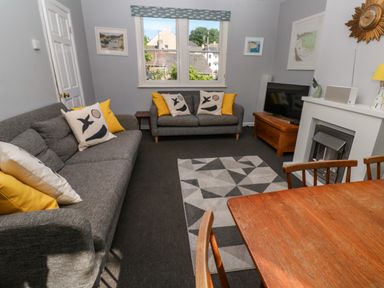 A living room with two gray sofas with yellow and patterned cushions a geometric rug a wooden dining table chairs a fireplace and a television at 8 Rosewall Cottages in St Ives