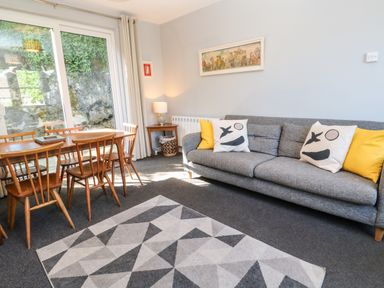 A living room with a gray sofa with printed cushions a wooden dining table with chairs and a geometric patterned rug at 8 Rosewall Cottages in St Ives