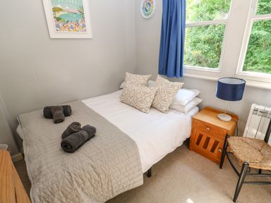 A bedroom with a bed with pillows and towels a wooden bedside table with a lamp a chair and a window with blue curtains at 8 Rosewall Cottages in St Ives
