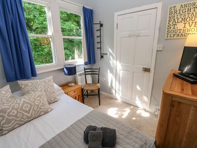 A bedroom with a bed pillows and towels a wooden nightstand with a lamp a chair a white door a wooden dresser with a television and a window with blue curtains at 8 Rosewall Cottages St Ives