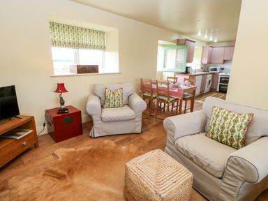 A living room with two armchairs a wicker ottoman a red side table with a lamp a wooden TV stand and a dining table with chairs and pink tablecloth at The Granary in Longwitton near Morpeth