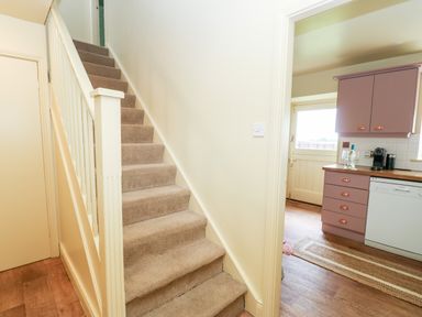 A hallway with carpeted stairs and a view into a kitchen with pink cabinets and wood flooring at The Granary in Longwitton near Morpeth