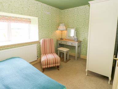 A bedroom with a striped armchair a vanity table with mirror and lamp a wardrobe and a bed with a blue cover at The Granary in Longwitton near Morpeth
