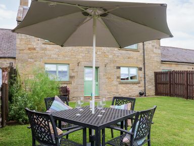 A garden with a black metal table and chairs under a large umbrella in front of a stone house with green windows and door at The Granary in Longwitton near Morpeth