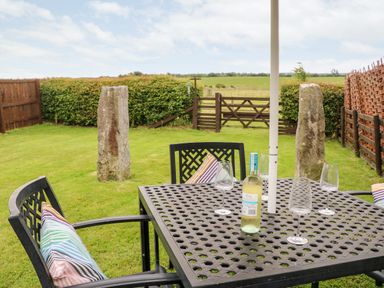 An outdoor patio table with four wine glasses and a bottle of white wine surrounded by chairs with striped cushions in a grassy garden with stone pillars and a wooden fence at The Granary in Longwitton near Morpeth