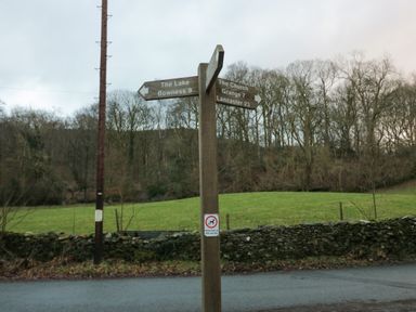 A countryside road signpost with directions to The Lake Bowness Grange and Lancaster by a field and forest