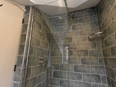 A corner shower area with green tiled walls a rainfall showerhead a handheld shower and a metal corner shelf at The Lodge in Newby Bridge