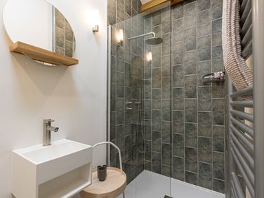 A bathroom with a shower area tiled in rectangular tiles a white sink with a faucet a round mirror with a wooden shelf and a gray towel on a radiator at The Lodge in Newby Bridge