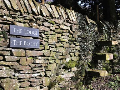 A stone wall with moss and ivy growing on it with two signs saying The Lodge and The Bothy at The Lodge in Newby Bridge