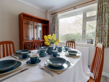 A dining room with a table set with blue dishes and cups yellow flowers in a vase wooden chairs and a glass cabinet by a window with floral curtains at Woodstock in Chaffcombe near Chard