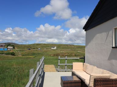 An outdoor patio with a cushioned sofa and table overlooking grassy fields and hills with wind turbines at Eala Bhan Cottage in Clachan near Balivanich