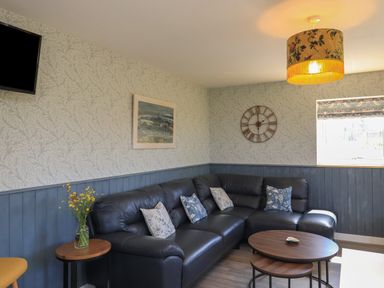 A living room with a black sectional sofa pillows round wooden coffee tables a wall clock a wall painting and a window at Eala Bhan Cottage Clachan near Balivanich