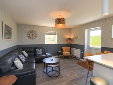 A living room with a black sectional sofa and cushions a round wooden coffee table a yellow armchair a wall clock and a door at Eala Bhan Cottage in Clachan near Balivanich