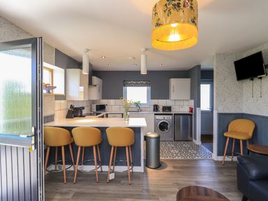 A kitchen with a breakfast bar and yellow stools next to a living area with a mounted TV at Eala Bhan Cottage in Clachan near Balivanich