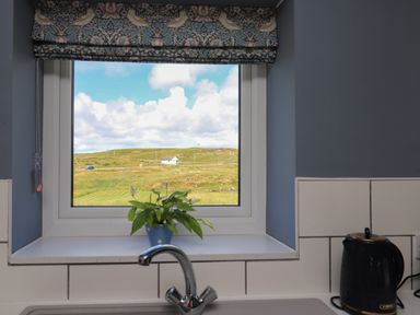 A kitchen window with a plant on the sill and a kettle on the counter at Eala Bhan Cottage in Clachan near Balivanich