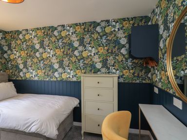 A bedroom with a bed a white chest of drawers a yellow chair a narrow white desk and floral wallpaper at Eala Bhan Cottage in Clachan near Balivanich