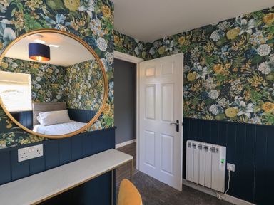 A room with floral wallpaper a white door a circular mirror reflecting a bed and window at Eala Bhan Cottage in Clachan near Balivanich