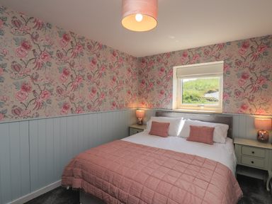 A bedroom with a double bed with pink bedding and floral wallpaper at Eala Bhan Cottage in Clachan near Balivanich