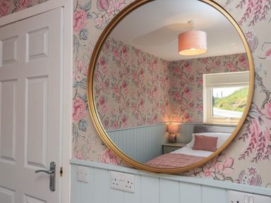 A bedroom with floral wallpaper a bed with a pink pillow and blanket a nightstand with a lamp and a window reflected in a round mirror at Eala Bhan Cottage in Clachan near Balivanich