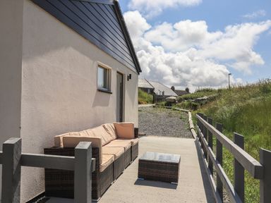 An outdoor patio with a cushioned wicker sofa and a small wicker table next to a building with a gravel path and grassy slope at Eala Bhan Cottage in Clachan near Balivanich