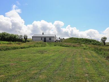 A grassy field with a white building in the background under a cloudy sky at Eala Bhan Cottage in Clachan near Balivanich