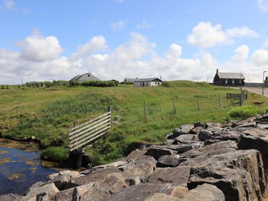 A rural landscape with grass fields a wooden gate a rocky embankment and houses in the distance at Eala Bhan Cottage in Clachan near Balivanich