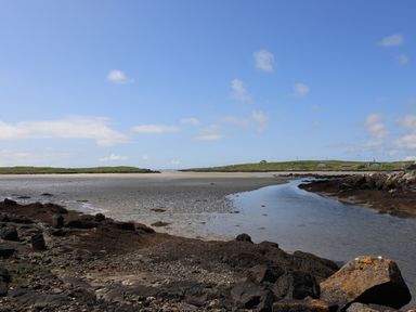 A coastal landscape with rocks and tidal water with green hills and small buildings in the background at Eala Bhan Cottage in Clachan near Balivanich