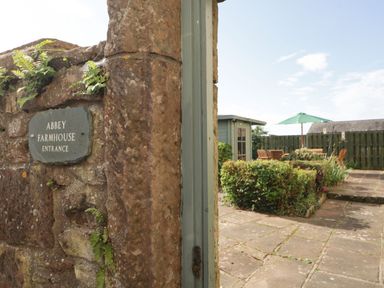 A stone entrance wall with a sign reading Abbey Farmhouse Entrance and a garden patio with chairs and a green umbrella at Abbey Farm House in St Bees