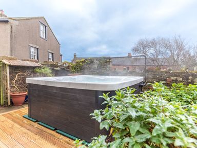 An outdoor hot tub on a wooden deck next to a stone wall with plants at Abbey Farm House in St Bees