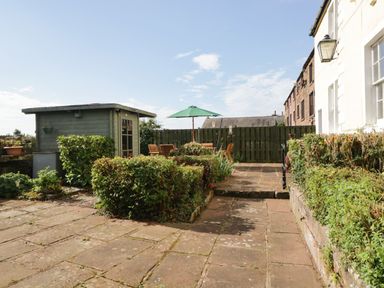 An outdoor patio area with stone paving green shrubs a wooden shed wooden table with chairs and a green umbrella at Abbey Farm House in St Bees