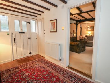 An entryway with a patterned rug a white panel door a radiator and a view into a living room with sofas and exposed ceiling beams at Poppy Cottage in Beaminster