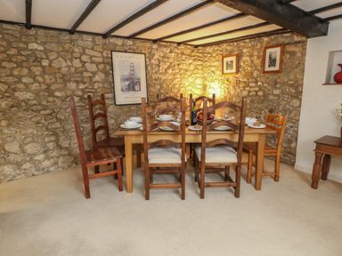 A dining room with a wooden table set for six with stone walls and framed pictures at Poppy Cottage in Beaminster