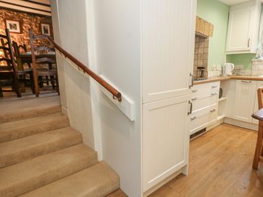 Carpeted stairs with wooden handrail leading to a dining area and adjacent kitchen with white cabinets and wooden floor at Poppy Cottage in Beaminster