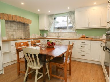 A kitchen with a wooden dining table and mixed chairs a bowl of fruit on the table white cabinets and a window with curtains at Poppy Cottage in Beaminster