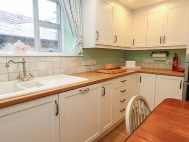 A kitchen with white cabinets wooden countertop a double sink a loaf of bread on a cutting board a fire extinguisher and a wooden dining table at Poppy Cottage in Beaminster