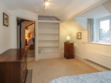 A bedroom with a wooden dresser a bedside cabinet with a lamp empty shelves and a window with floral curtains at Poppy Cottage in Beaminster